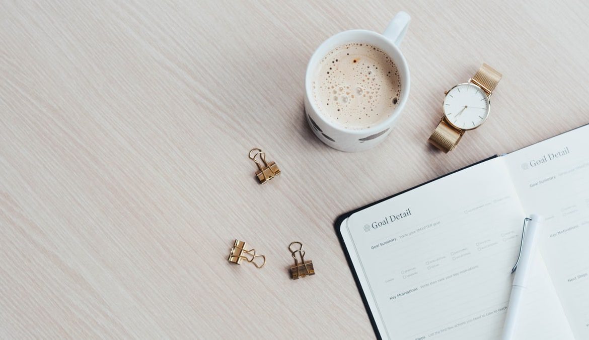 A minimalist desk arrangement featuring a coffee cup, notebook, pen, and wristwatch.
