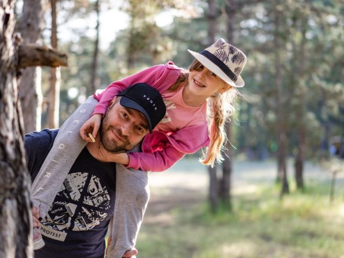 A joyful father giving his daughter a piggyback ride in a sunny forest setting.